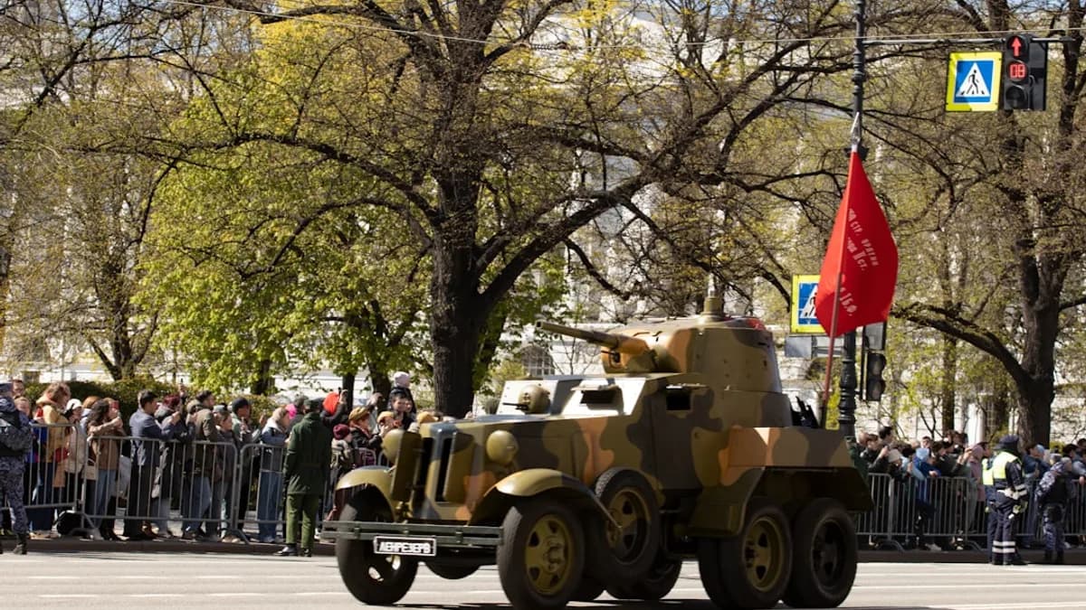 A camouflaged armored car drives through a crowd.