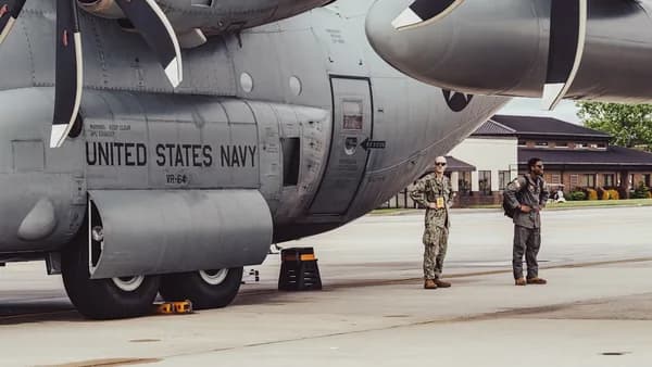 Two navy personnel stand by a military aircraft.