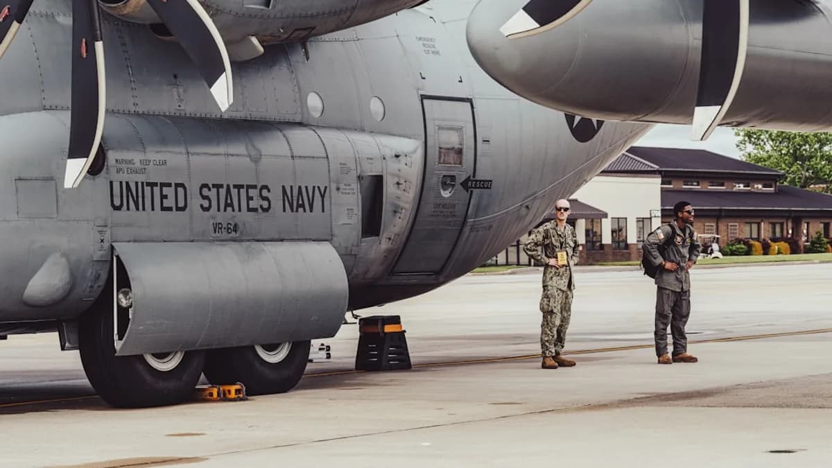 Two navy personnel stand by a military aircraft.