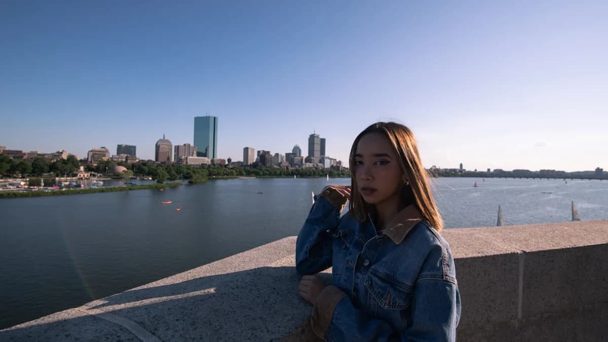 woman in blue denim jacket sitting on gray concrete pavement near body of water during daytime