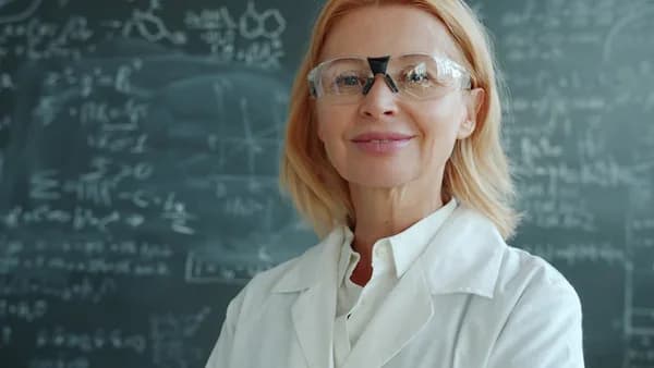 Woman in lab coat and glasses in front of blackboard.